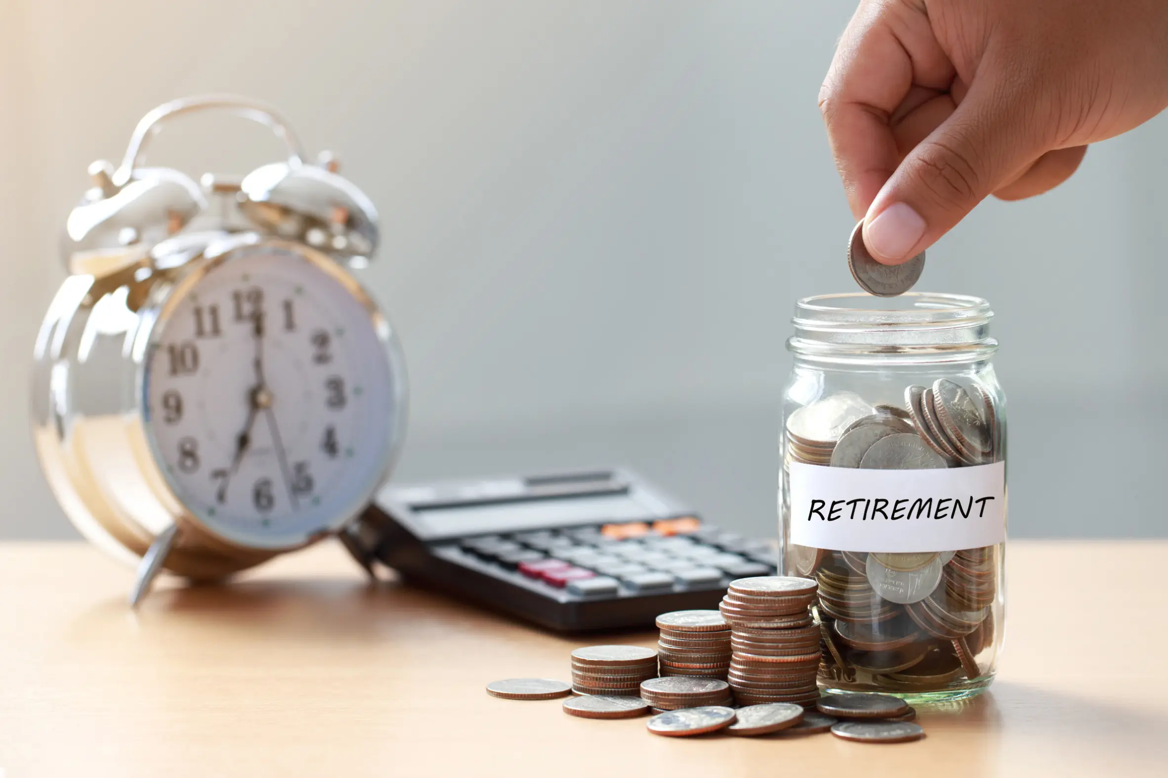 Hand placing a coin into a retirement savings jar with calculator and clock representing financial planning